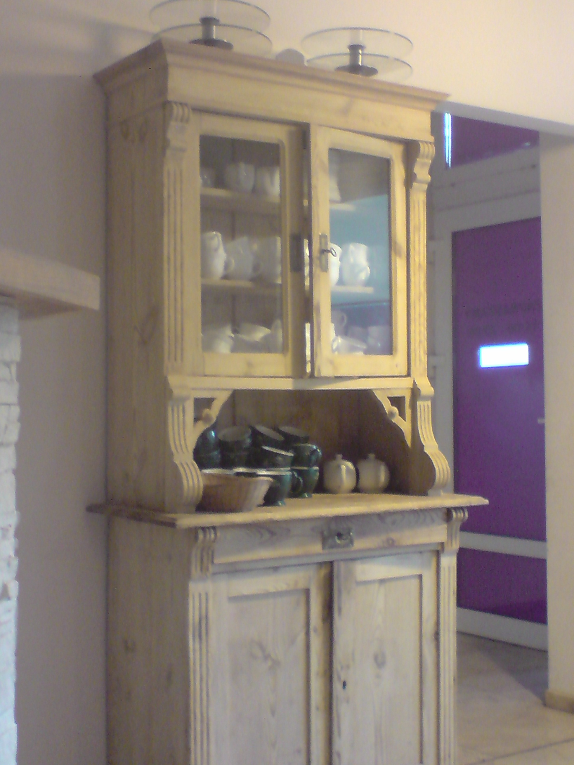 Drewniany kredens w stylu retro z przeszkloną górną częścią, eksponujący białą porcelanę. Na blacie ustawione ceramiczne naczynia i dekoracyjne dynie. W tle widoczna fioletowa ściana.